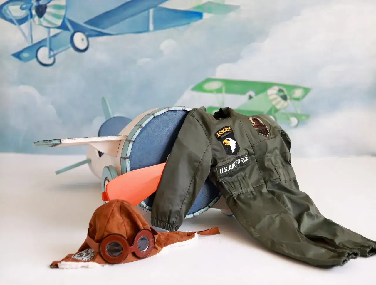 Stroj pilota dla dzieci z naszywkami U.S. AIRFORCE i AIRBORNE, obok drewniany samolot i czapka z goglami.