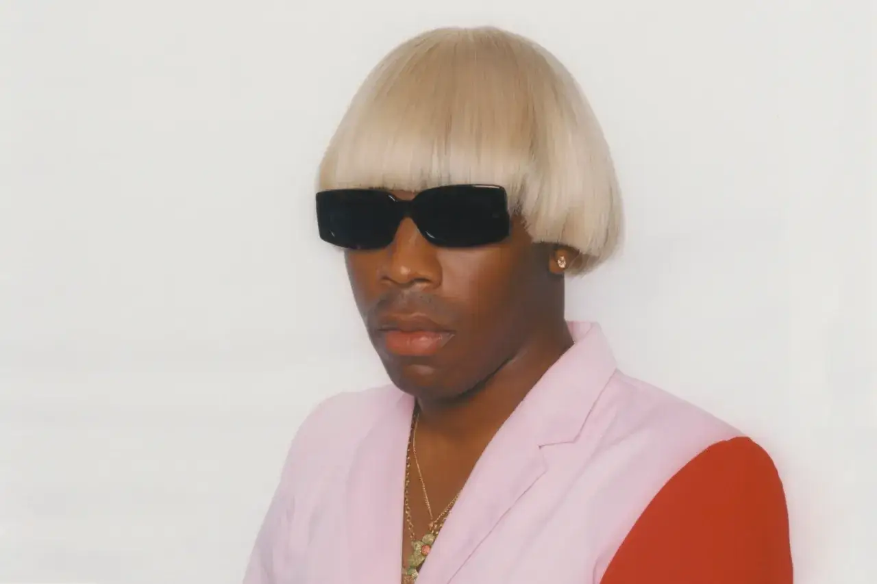 Igor Tyler the Creator w peruce i okularach przeciwsłonecznych, ubrany w różową marynarkę z czerwonym rękawem.
