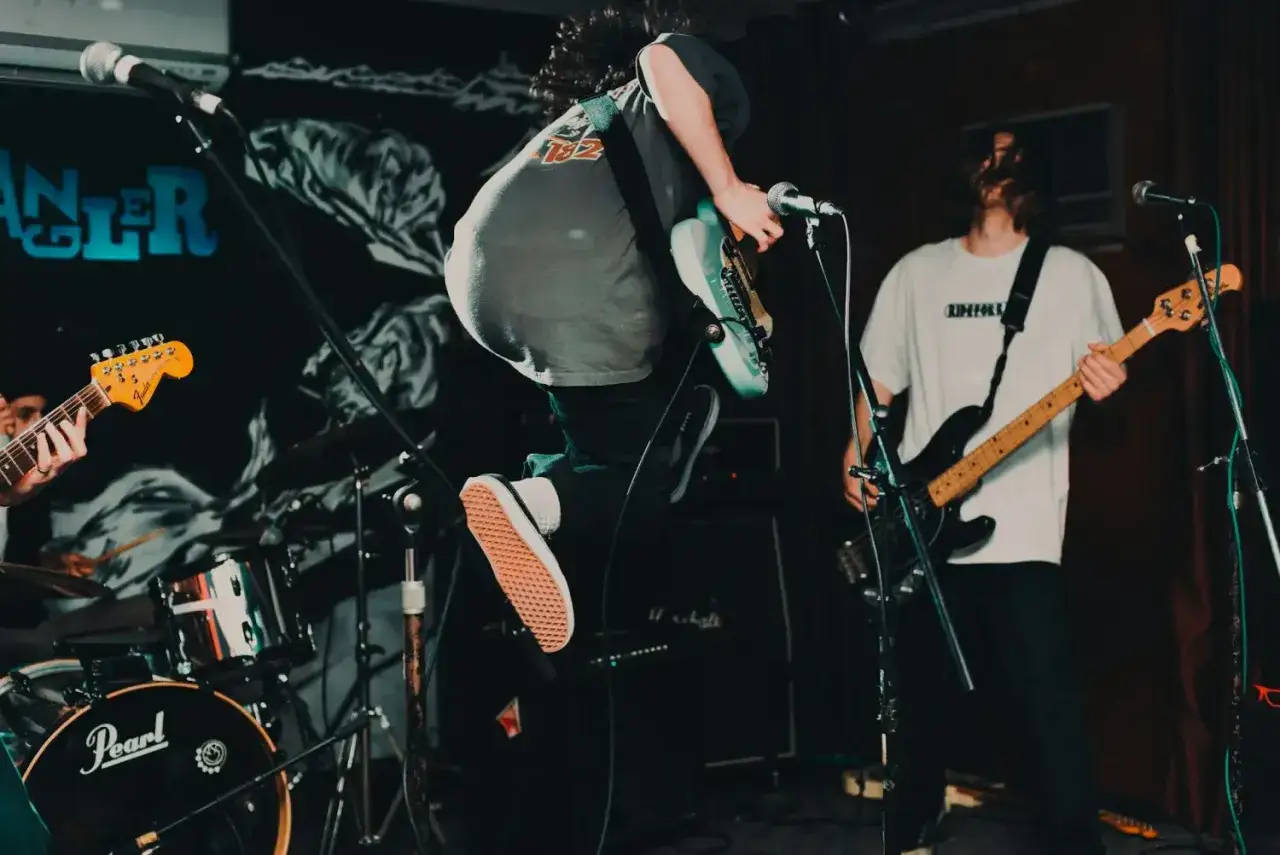 Ein Gitarrist springt auf der Bühne, ein Bassist spielt im Hintergrund. Energiegeladener Auftritt im Stil des **punk 70er**.
