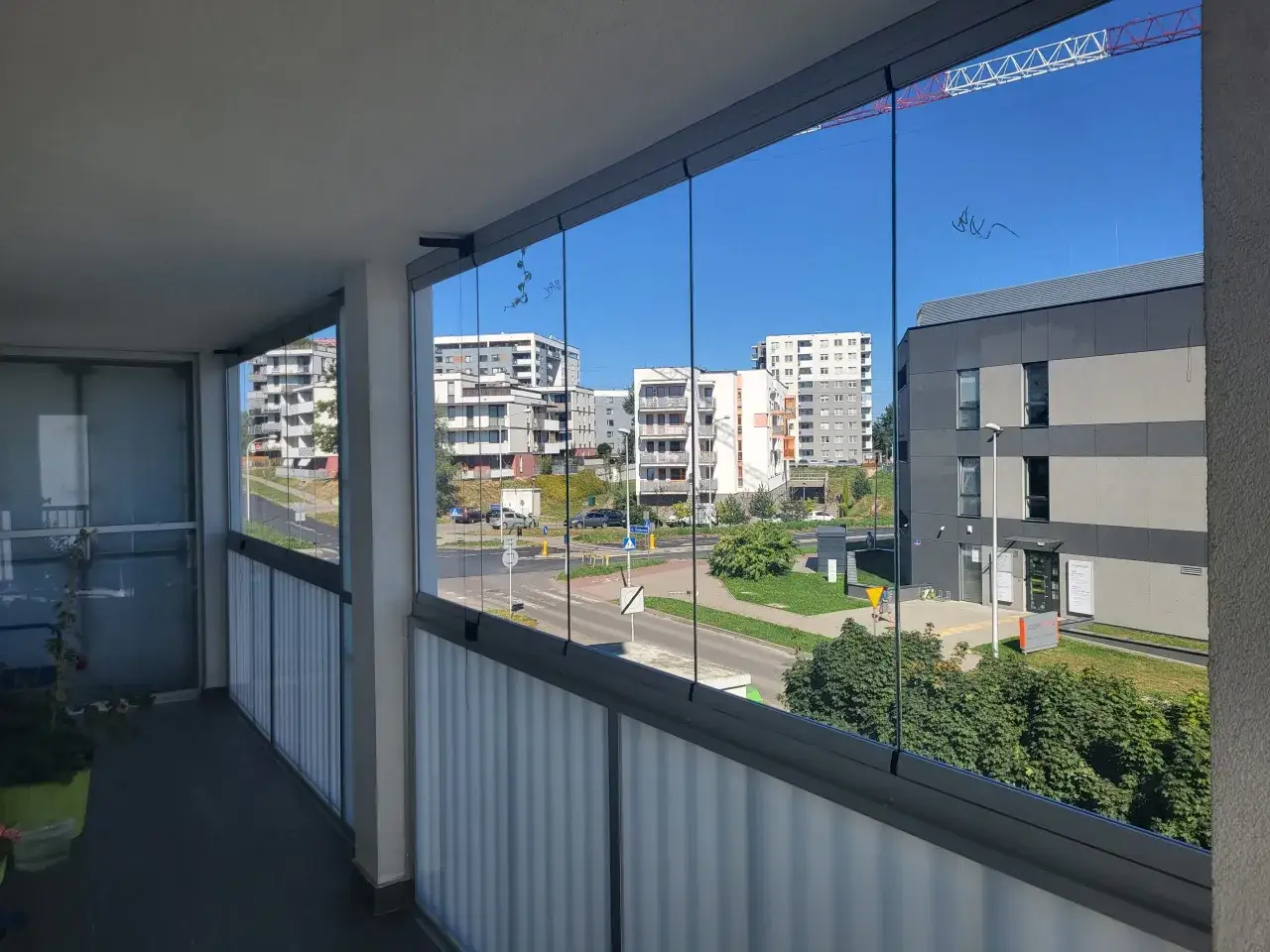 Zabudowa loggi w bloku z widokiem na osiedle i plac budowy. Szklane panele chronią przed wiatrem, tworząc dodatkową przestrzeń.