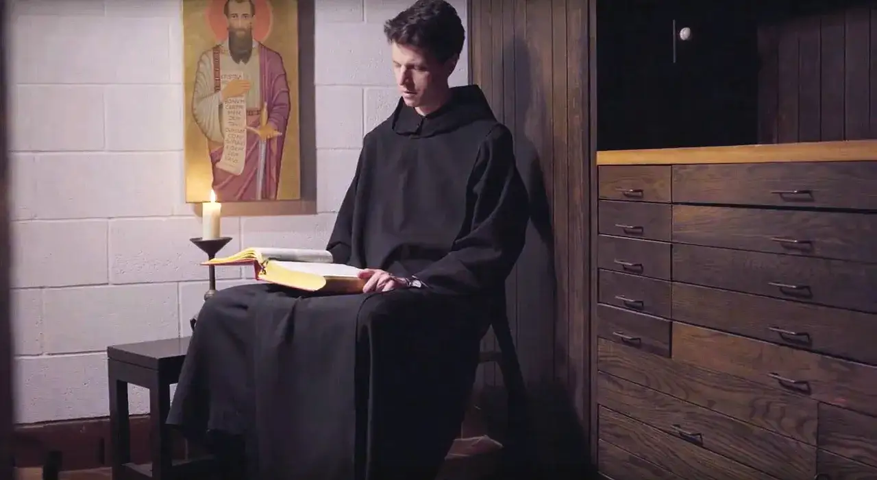 A monk in a black habit reads a book by candlelight, a painting of a saint behind him. This scene reflects the essence of the st. benedict rules summary.