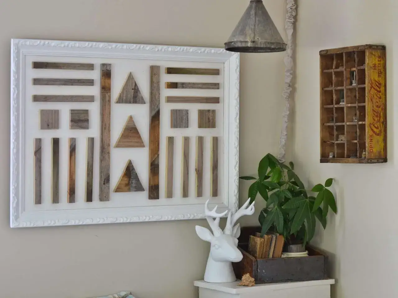 Une oeuvre d'art maison en bois recyclé, composée de formes géométriques, encadrée de blanc. Une tête de cerf et une plante complètent la scène.