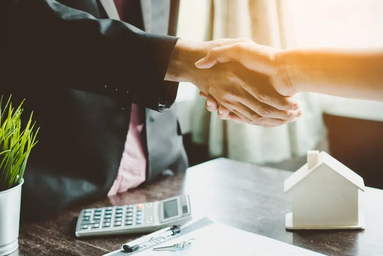 Poignée de main entre deux personnes, symbolisant un accord. Une calculatrice, des clés et une maquette de maison sont sur la table, suggérant une transaction immobilière. CCMI def.