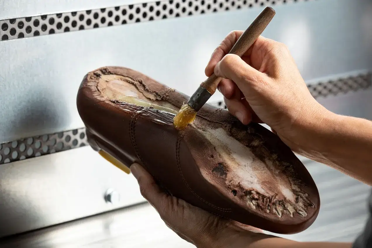 Main d'artisan appliquant de la colle sur une semelle de chaussure marron pour la réparer. L'artisan utilise un pinceau pour **poser soi même patin chaussure**.