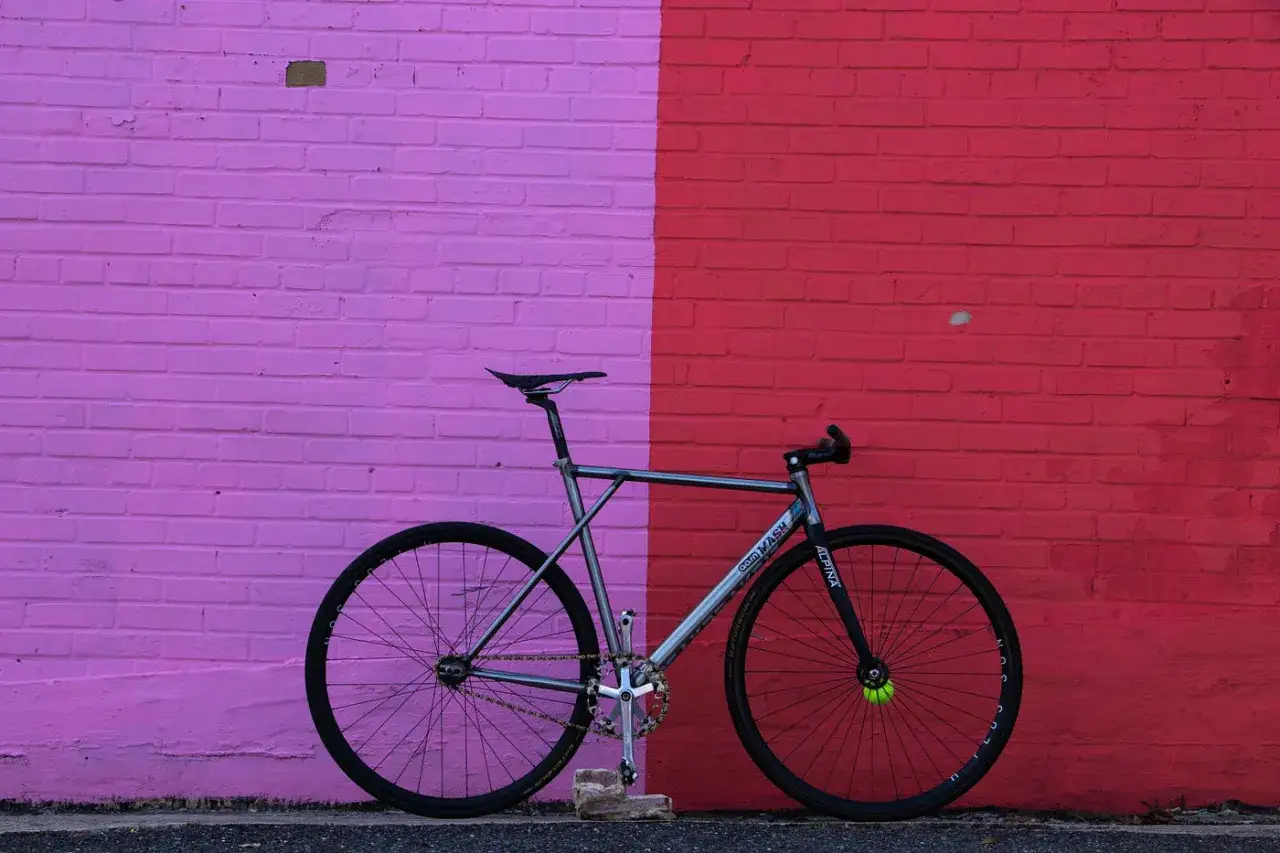 A sleek silver fixed gear bike, possibly a MASH brand, leans against a vibrant pink and red brick wall.