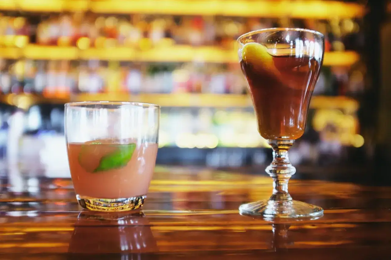 Due cocktail su un bancone di legno lucido, uno con una fetta di lime e l'altro con una guarnizione verde. Un classico **bourbon drink** pronto da gustare.