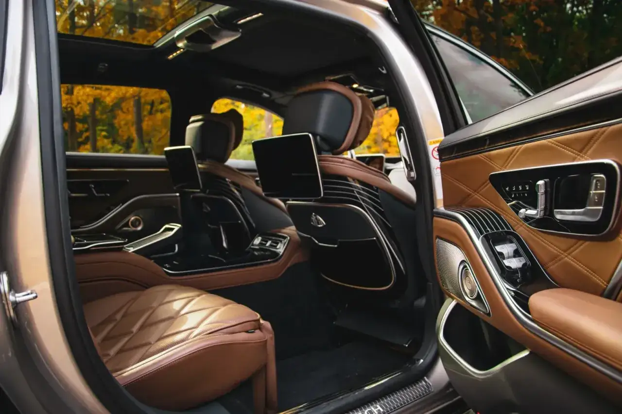 Luksusowe wnętrze Maybach S680 z brązową skórzaną tapicerką, ekranami dla pasażerów i jesiennym krajobrazem za oknem.