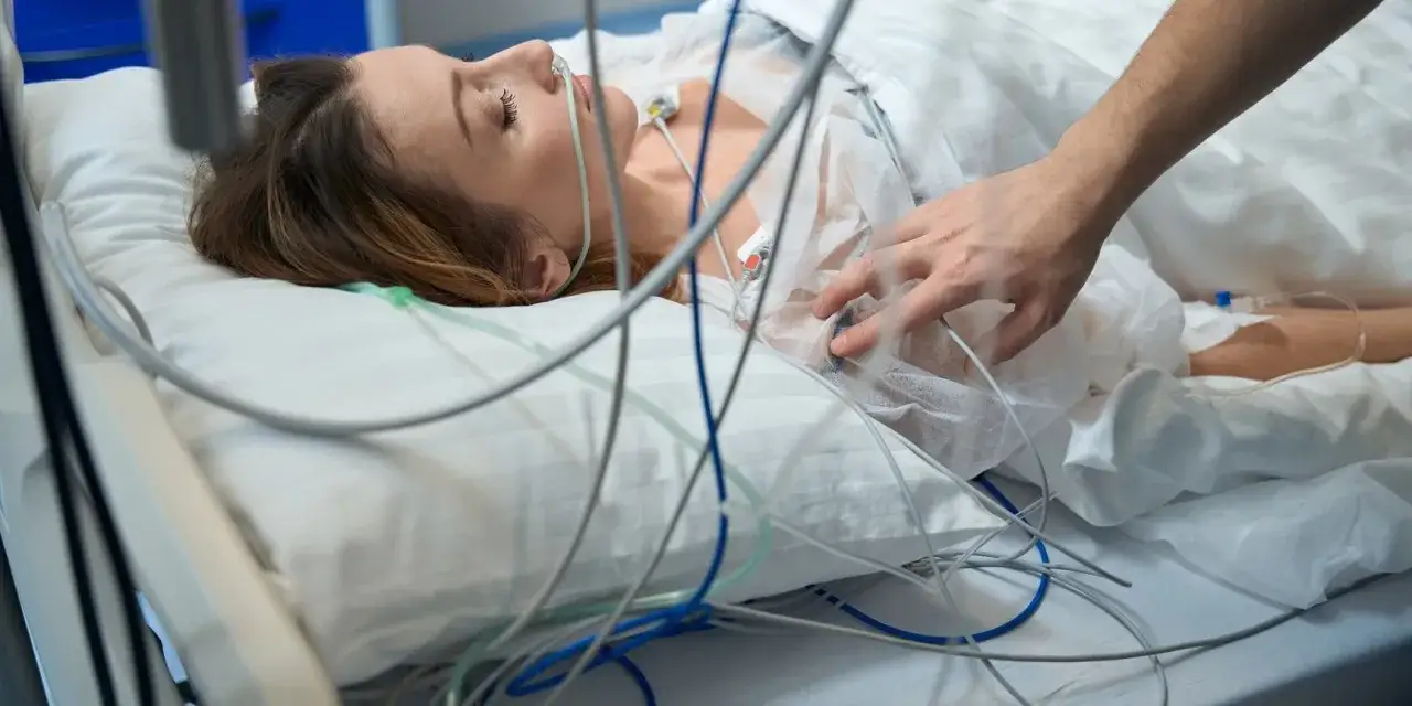 Kobieta leży podłączona do aparatury medycznej, z rurką tlenową w nosie. Dłoń bliskiej osoby delikatnie dotyka jej klatki piersiowej, łagodząc ból głowy po narkozie.