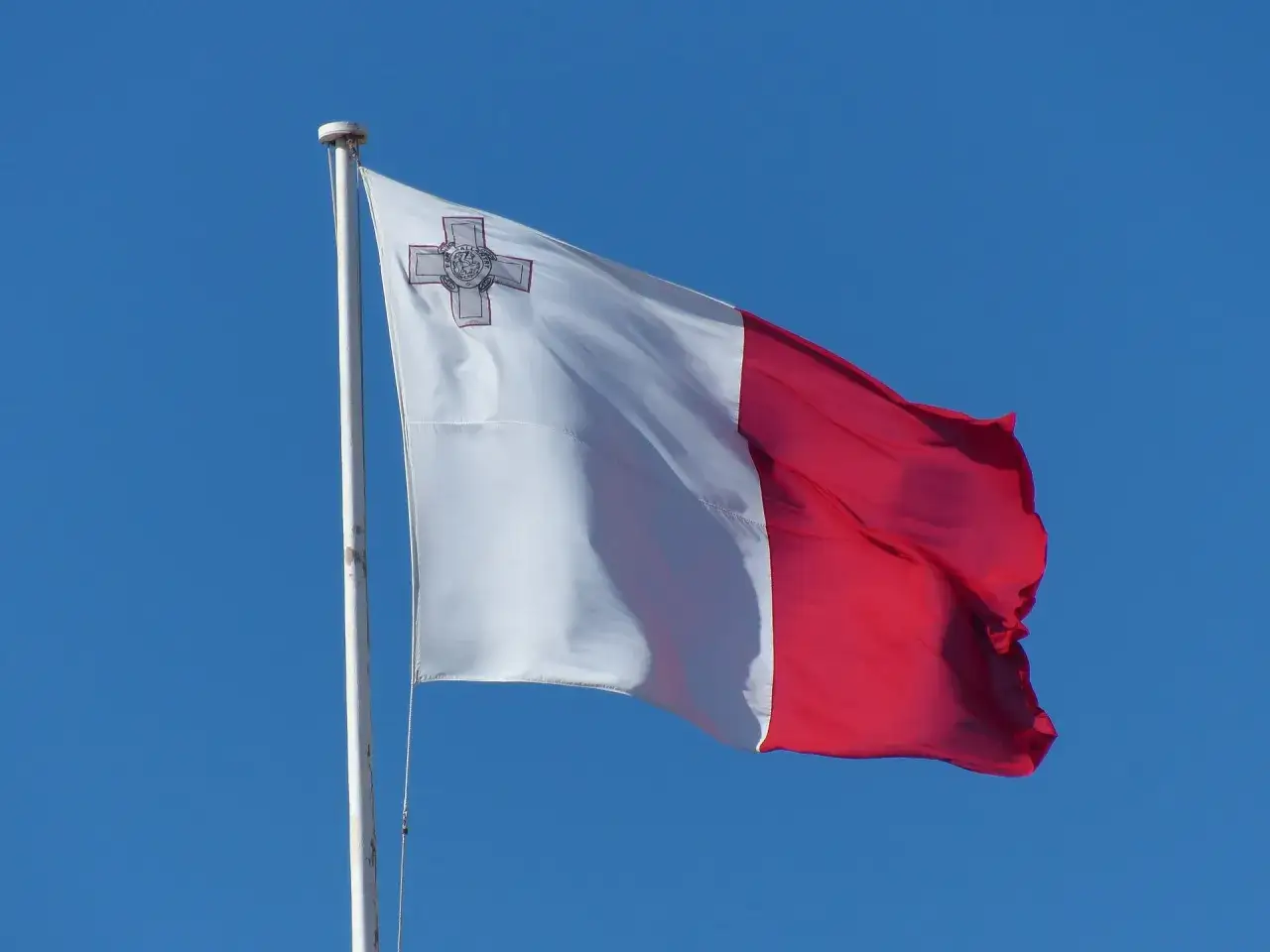 Flaga Malty powiewa na wietrze na tle błękitnego nieba. Biało-czerwona flaga z symbolem krzyża na białej części.