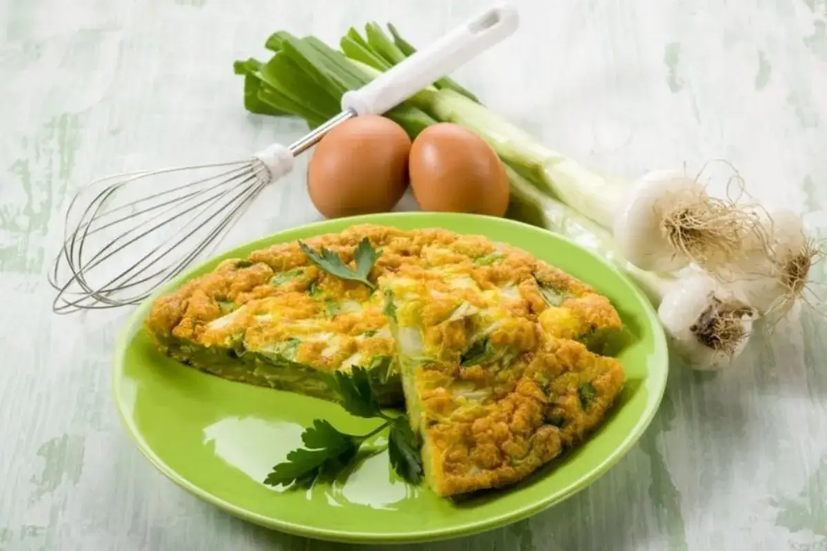 Frittata con cavolo nero, uova, cipollotti e frusta su un piatto verde.