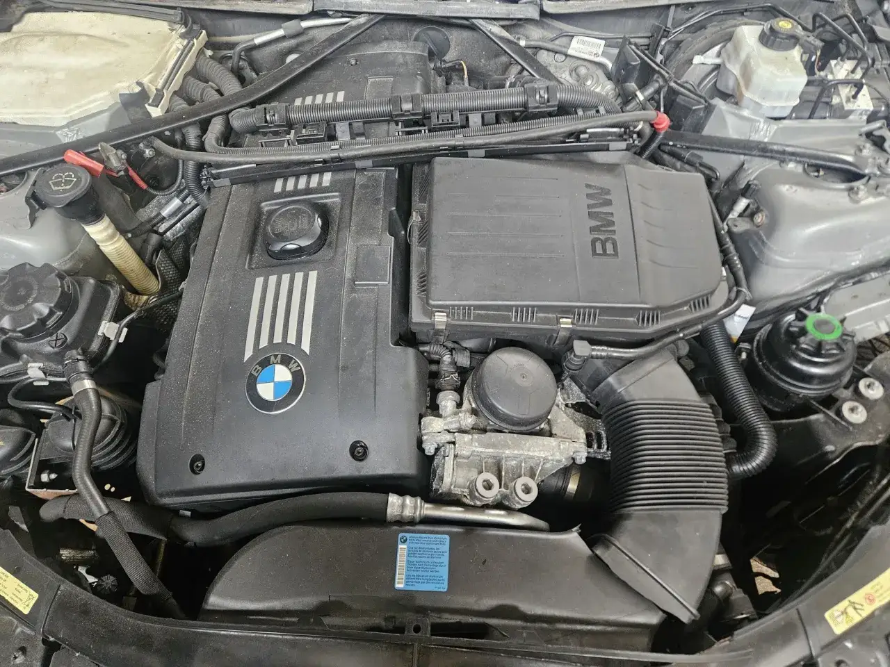Silnik BMW 335, widok z góry. Widoczne elementy osłony silnika z logo BMW, przewody i elementy układu dolotowego.