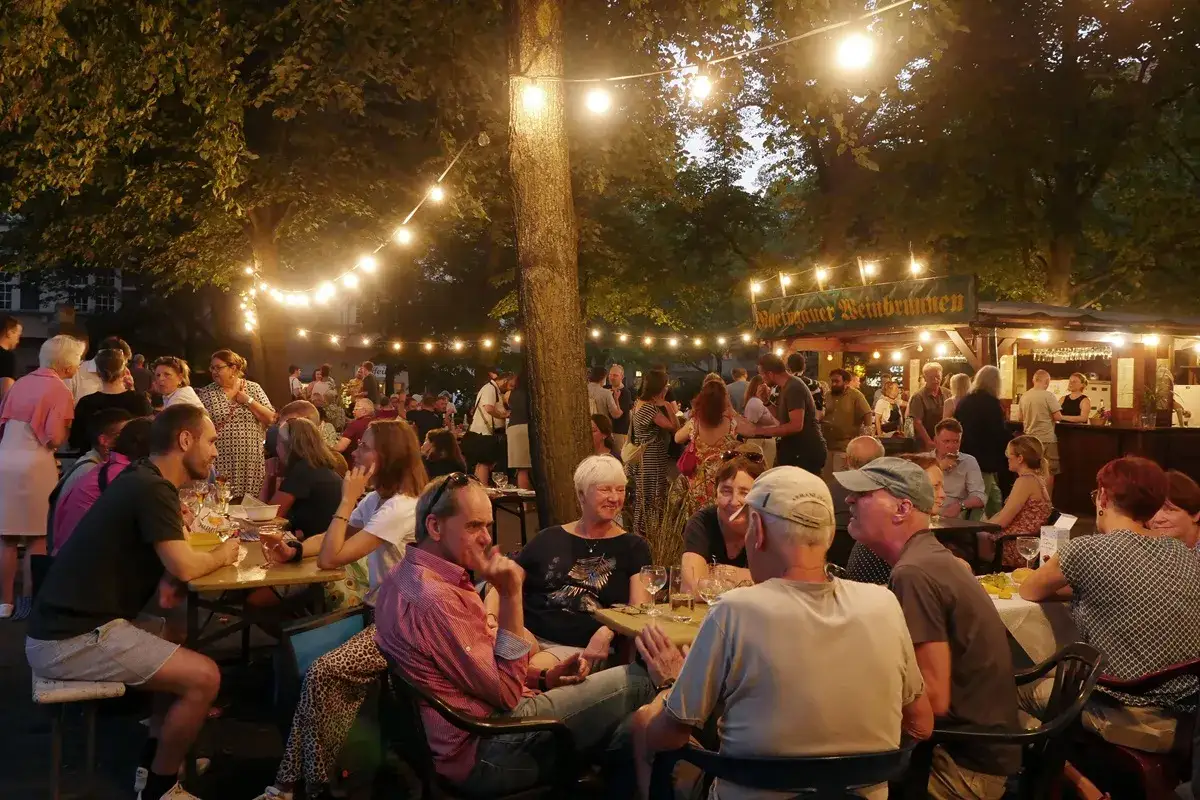 Menschen genie&szlig;en ein Weinfest in Berlin unter Lichterketten. Ein Weinbrunnen ist im Hintergrund zu sehen.