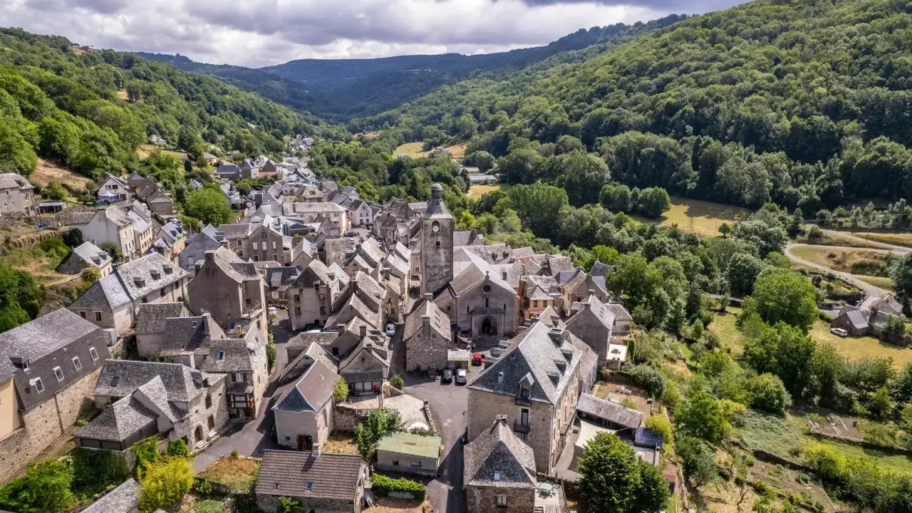 Vue aérienne d'un village pittoresque niché dans une vallée verdoyante, rappelant le charme de Aumont-Aubrac à Conques.