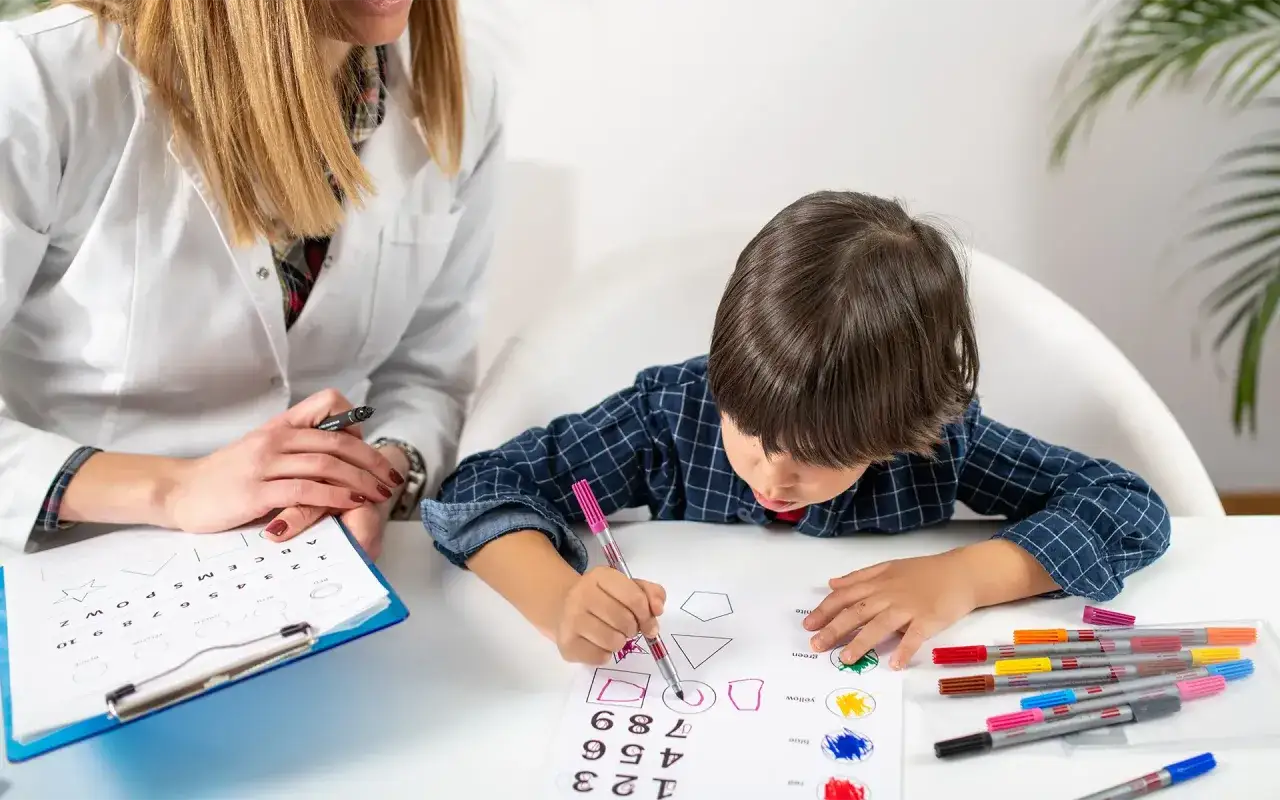 Un enfant concentré dessine des formes lors d'un test pour évaluer sa dyslexie. Une orthophoniste observe attentivement.
