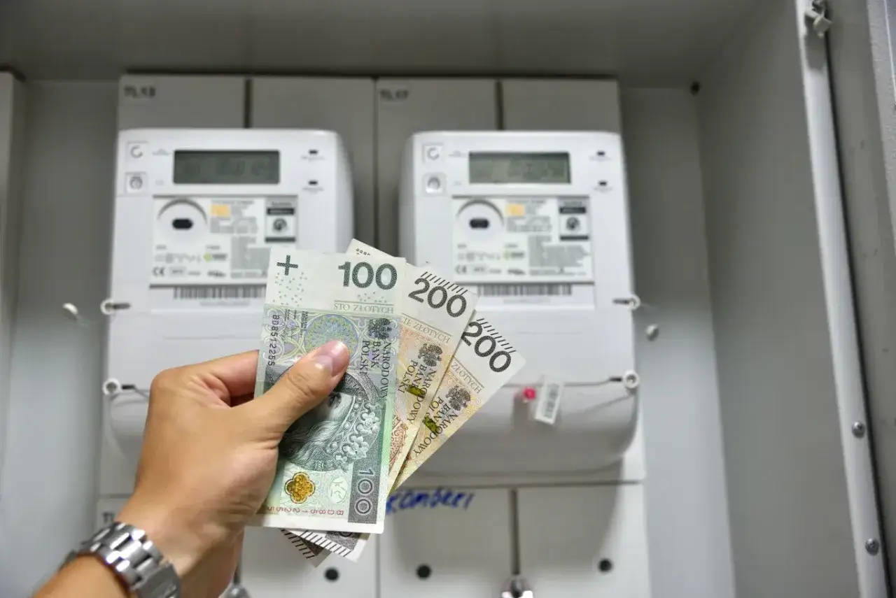 Dłoń trzyma polskie banknoty 100 i 200 zł przed licznikami energii elektrycznej. Czy to rachunek za prąd przy taryfie g12w?