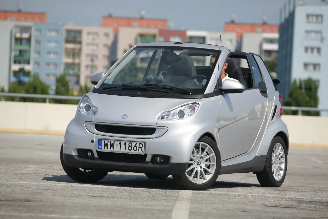Srebrny kabriolet Smart Fortwo 451 z otwartym dachem, z kierowcą w środku, na tle blok&oacute;w mieszkalnych.
