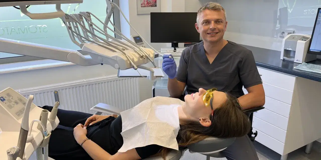 Dentysta w gabinecie z pacjentką na fotelu, trzymający narzędzie.