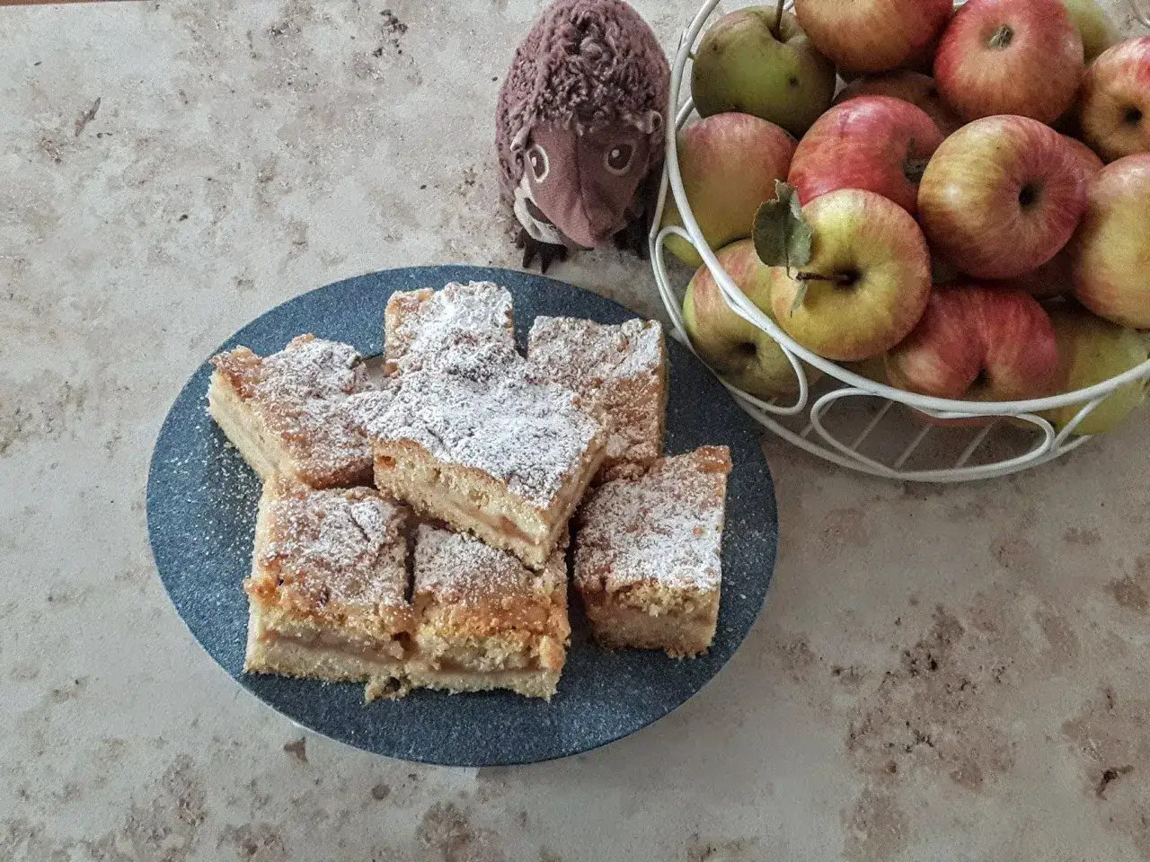 Szybka szarlotka z jabłkami ze słoika, posypana cukrem pudrem, obok koszyk jabłek i pluszowy jeż.