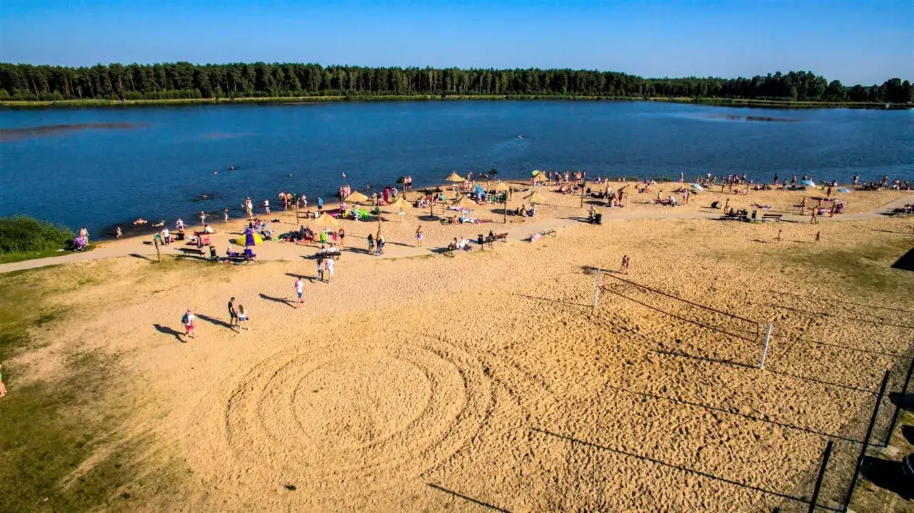 Plaża nad jeziorem, pełna ludzi opalających się i kąpiących. W oddali las, a na piasku boisko do siatkówki. To idealne miejsce na letni **wykrot**.