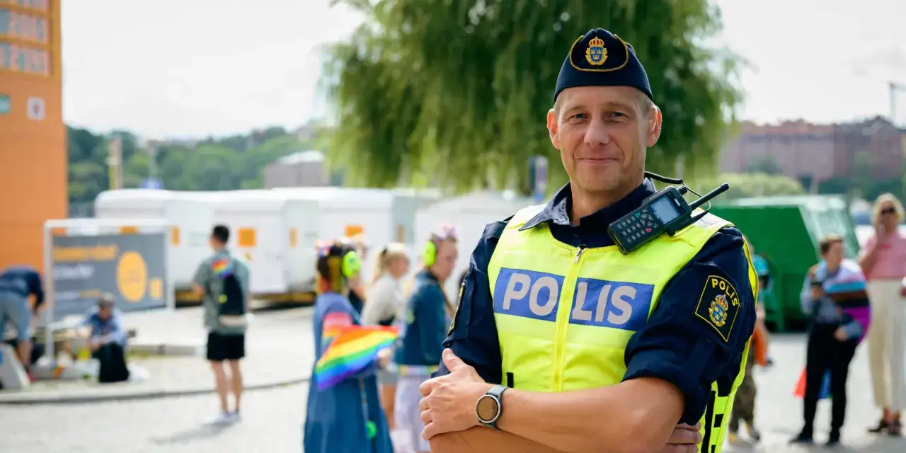 Polis i tj&auml;nst, kanske p&aring; v&auml;g att plugga till polis. I bakgrunden syns folk med regnb&aring;gsf&auml;rger.