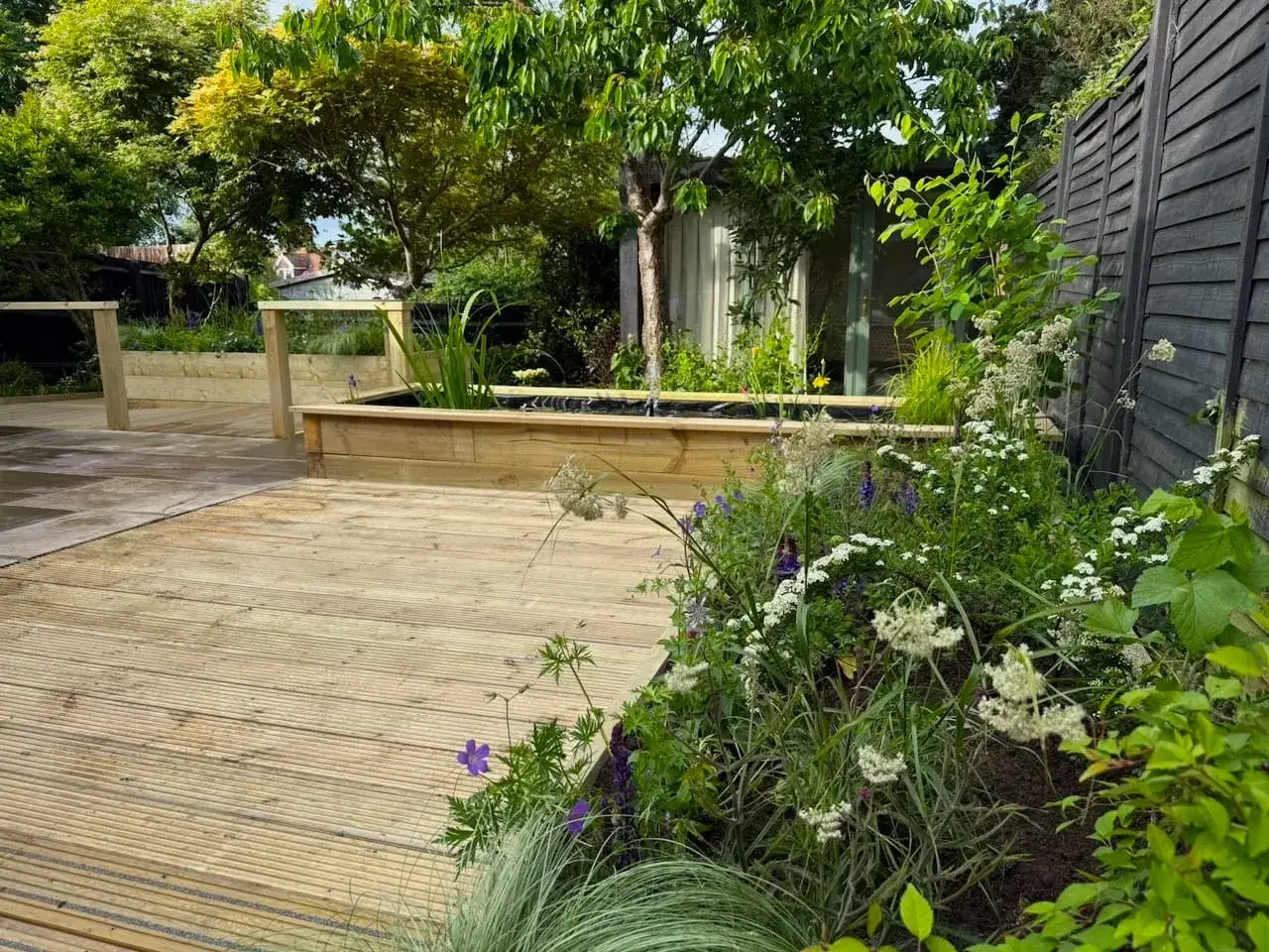 Moderner Garten am Hang mit Holzdeck, Teich und üppiger Bepflanzung.