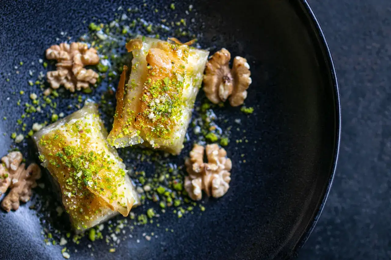 Zwei goldbraune Baklava-St&uuml;cke, bestreut mit gehackten Pistazien und Waln&uuml;ssen, auf einem dunklen Teller. Ein k&ouml;stliches Baklava Rezept.