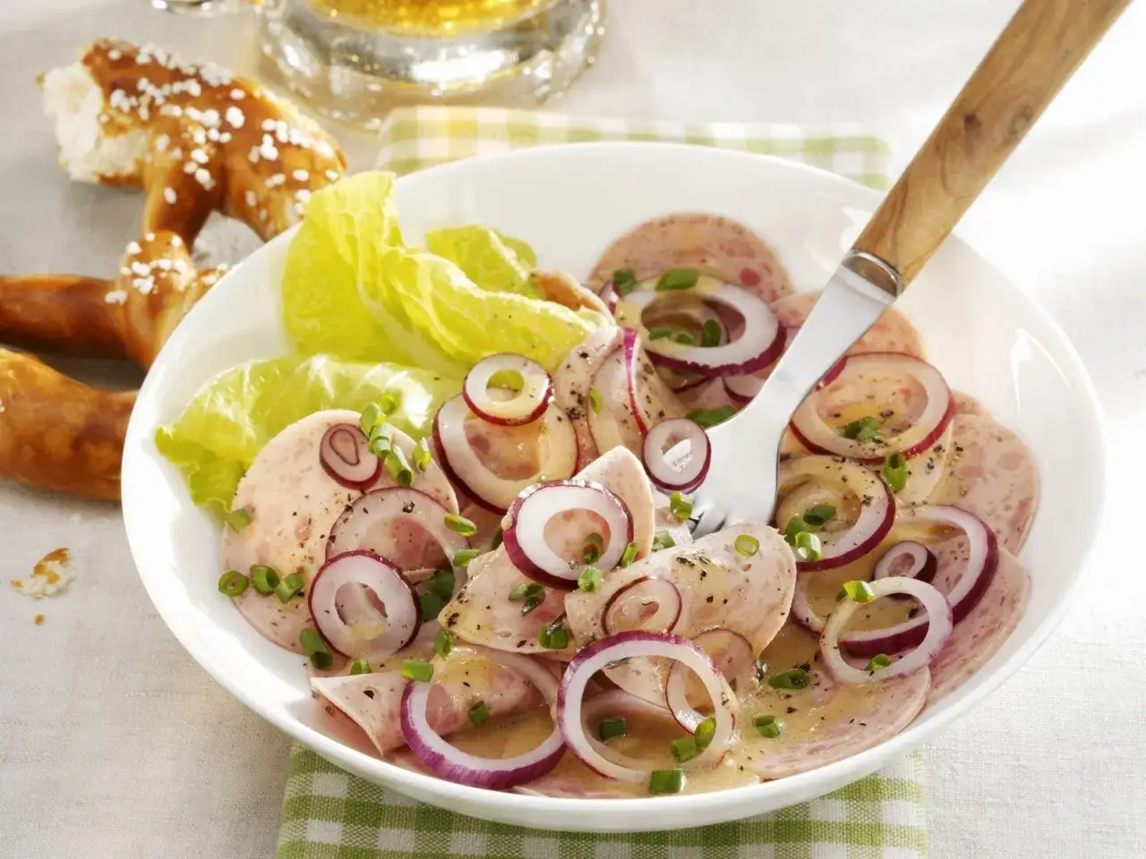 Ein herzhafter bayrischer Wurstsalat mit roten Zwiebelringen, Schnittlauch und einem Dressing, serviert mit Brezeln und Bier.