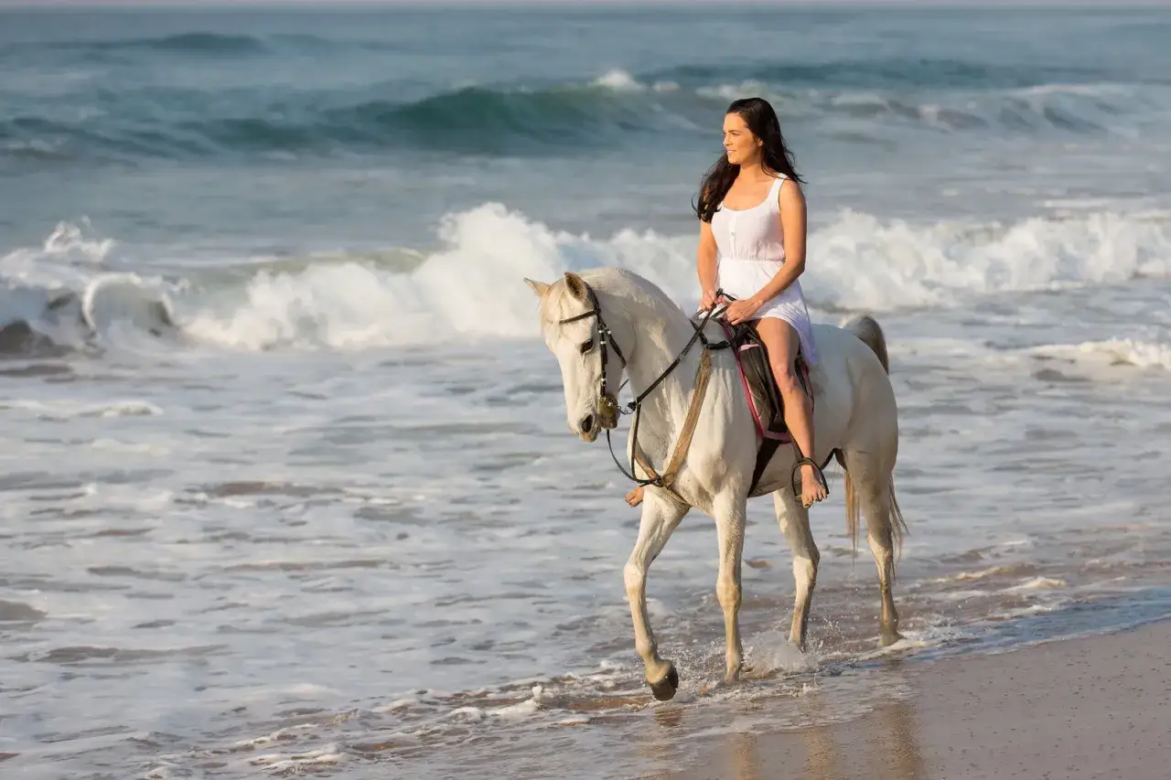 Kobieta w białej sukience na białym koniu, galop po plaży, fale rozbijają się o brzeg.