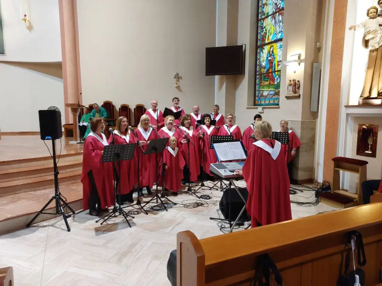 Schola w czerwonych szatach śpiewa w kościele. W tle widać witraż i ołtarz.