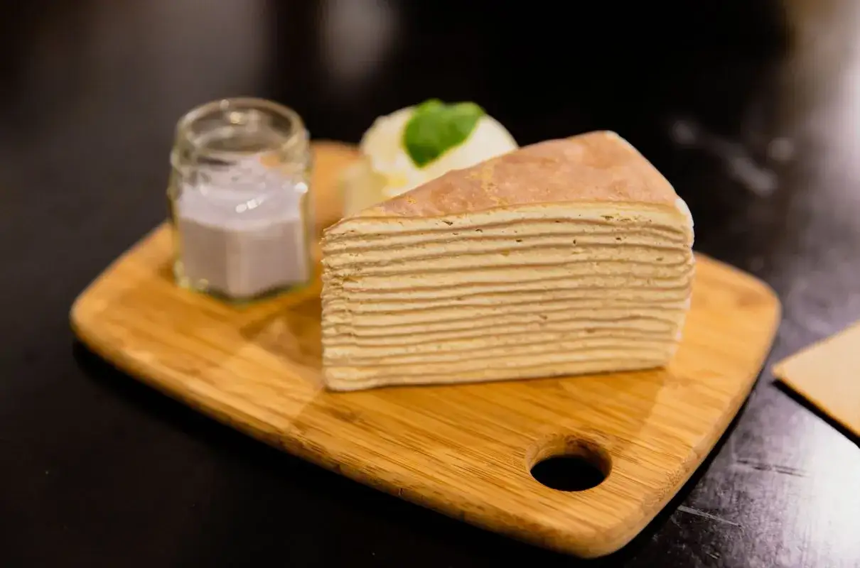 Kawałek ciasta mille-crêpe z kremem do przełożenia, podany z lodami i sosem w słoiczku na drewnianej desce.