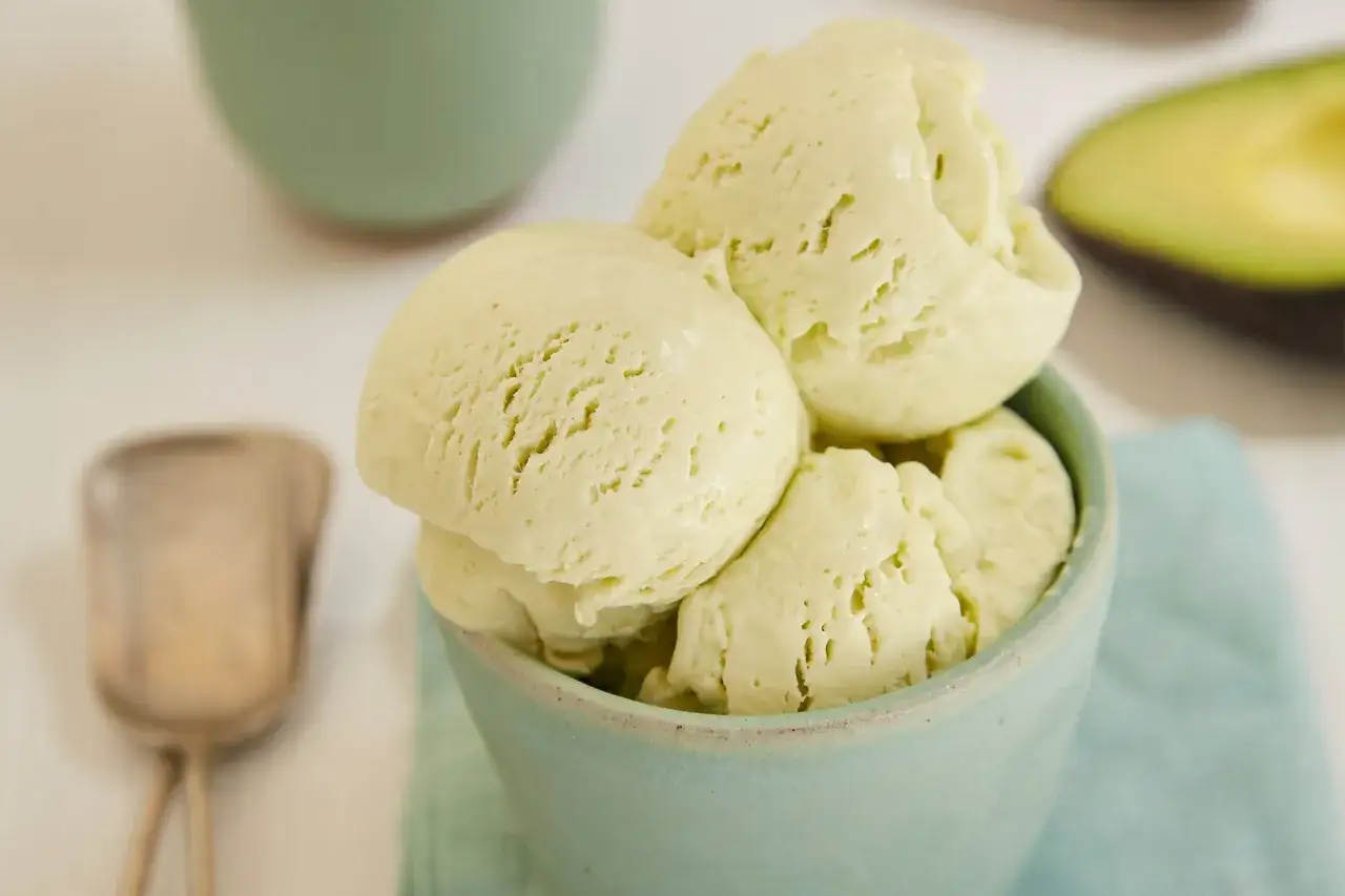 Un cremoso helado de aguacate en un cuenco azul claro, con una cuchara y medio aguacate al fondo.