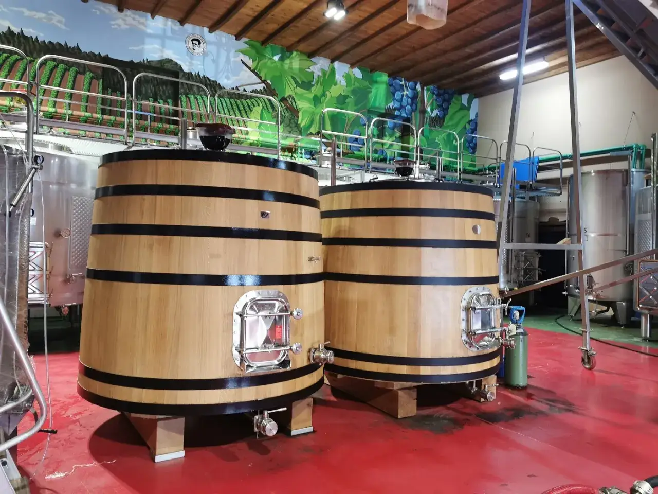 Dos grandes tinajas de madera, parte de un tratado de enología, reposan en una bodega. Detrás, murales de viñedos y racimos de uvas.