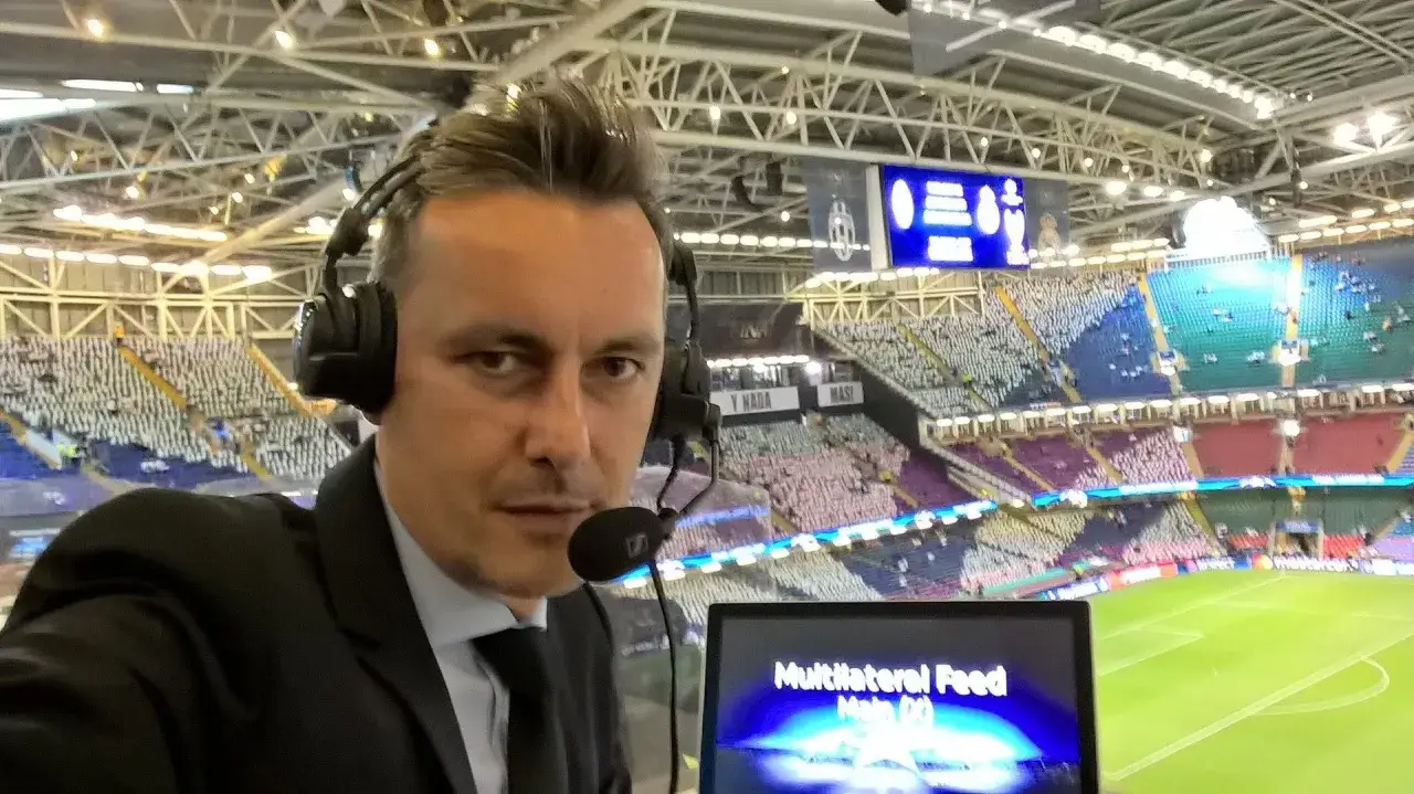 Rafał Wolski, komentator sportowy, w słuchawkach i z mikrofonem, na tle stadionu z widzami.
