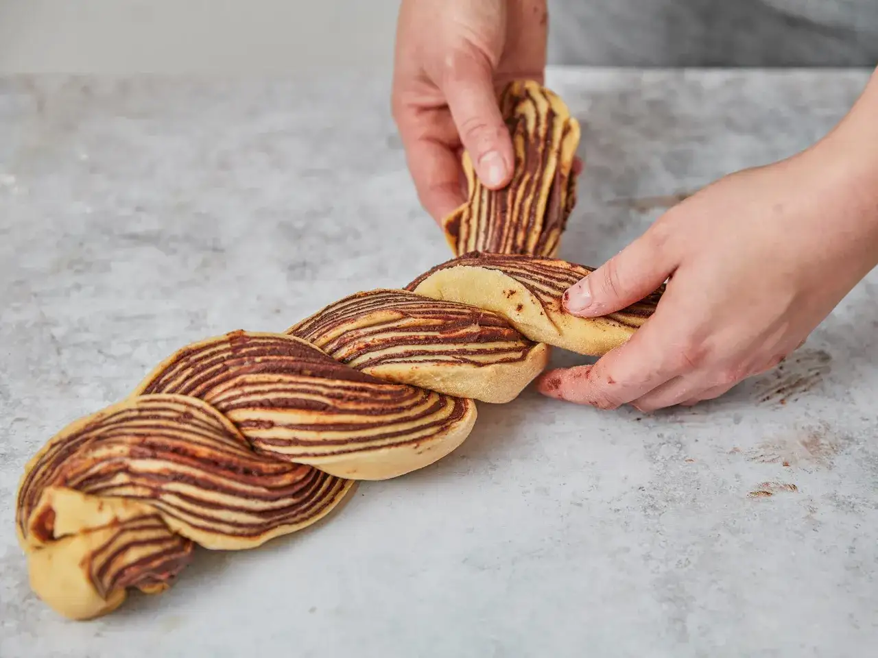 Hände flechten einen Teig mit Schokoladenstreifen, der an ein Pain au Chocolat erinnert.