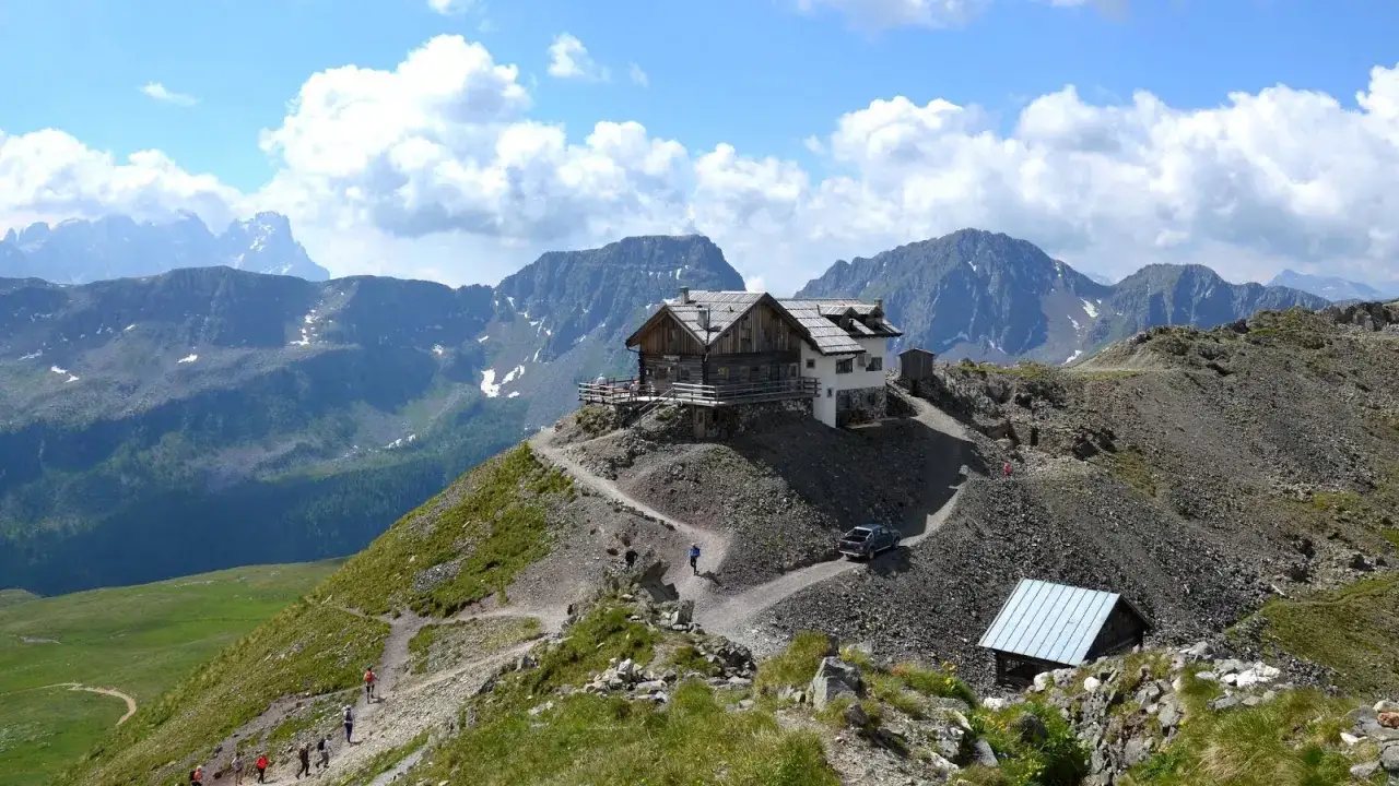 Rifugio alpino su un crinale roccioso, con sentiero che sale e persone che camminano. Sullo sfondo, montagne maestose e cielo azzurro con nuvole. Un'auto si avvicina al passo stalle.