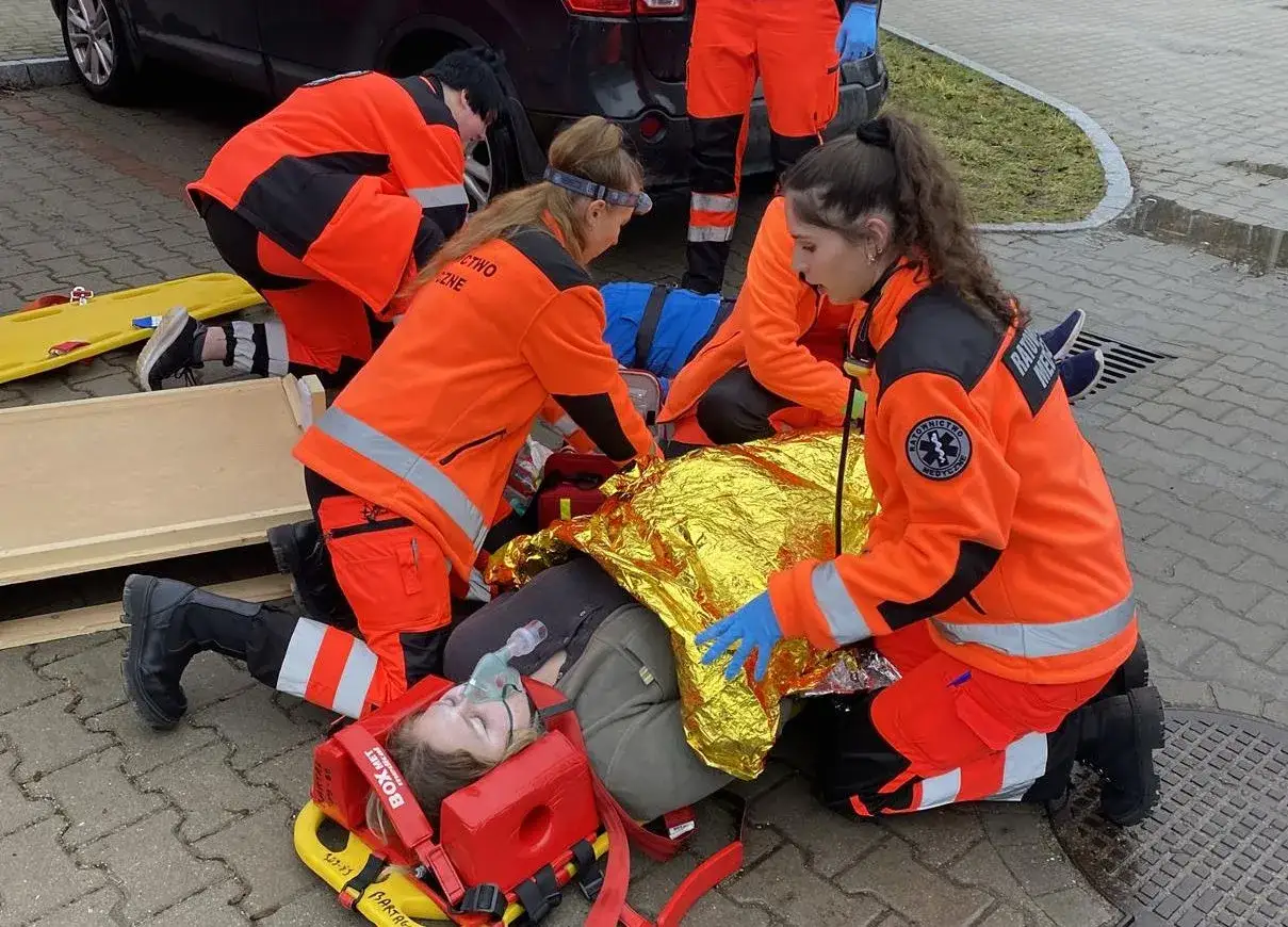 Uczestnicy kursu ratownika medycznego ćwiczą udzielanie pomocy osobie poszkodowanej, która jest podłączona do tlenu i okryta kocem termicznym.
