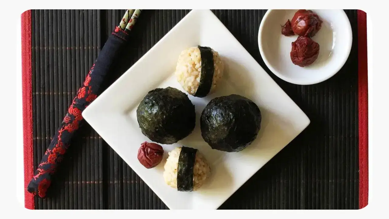 Cztery onigiri, dwa w całości owinięte w nori, dwa z widocznym ryżem i paskiem nori, obok miseczka z marynowanymi śliwkami ume.