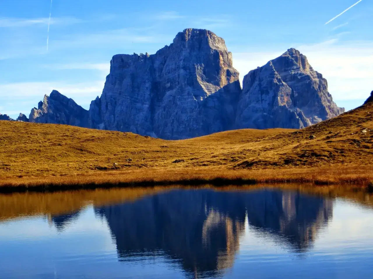 Riflessi maestosi delle Dolomiti nel lago delle Baste, con un prato dorato che separa le acque dalle imponenti vette.