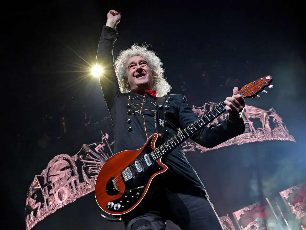 Brian May z zespołem Queen na scenie, z uniesioną pięścią i gitarą Red Special.