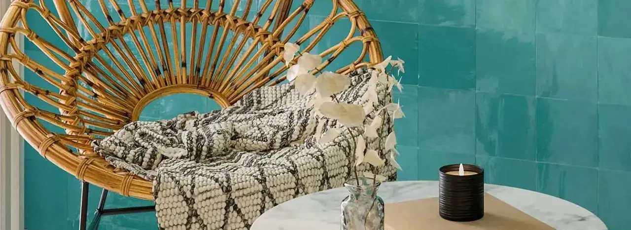 Chaise en rotin, plaid textur&eacute; et bougie sur une table, le tout devant un mur de carreaux bleus rappelant le zellige.