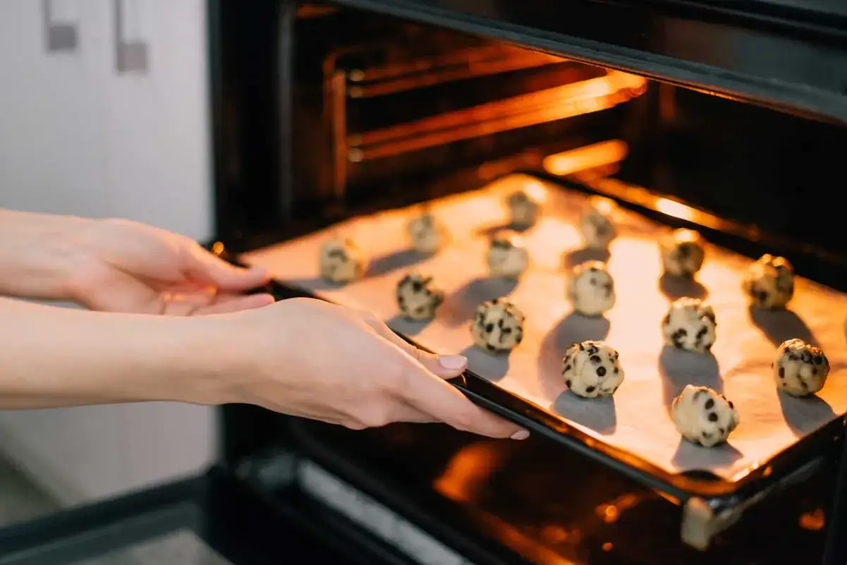 Manos colocando una bandeja con bolas de masa para galletas con chispas de chocolate en un horno caliente. &iexcl;As&iacute; es como hacer galletas caseras!
