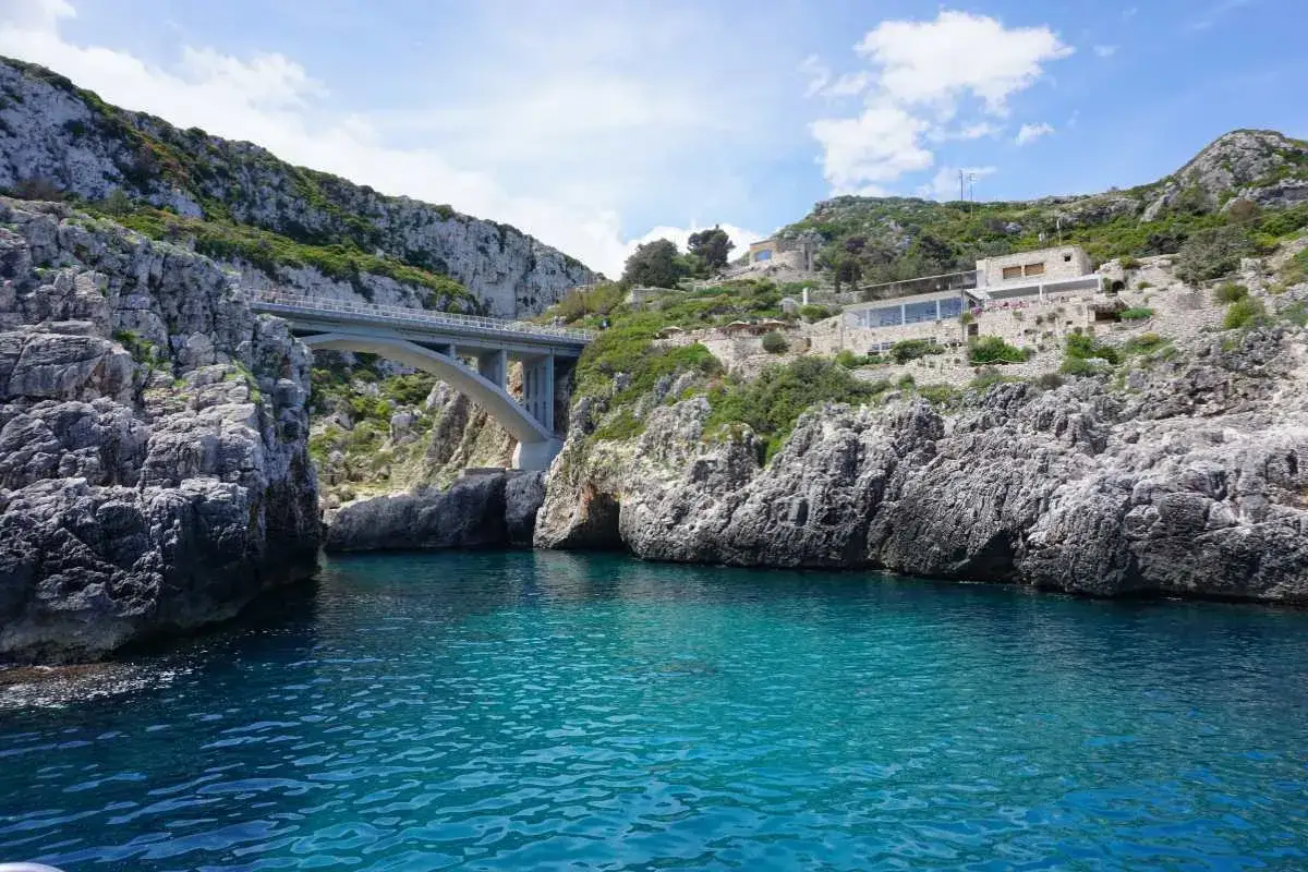 Ponte moderno su un mare cristallino, incastonato tra scogliere rocciose. Un luogo magico dove si incontrano i due mari in Puglia, con un ristorante panoramico sulla collina.