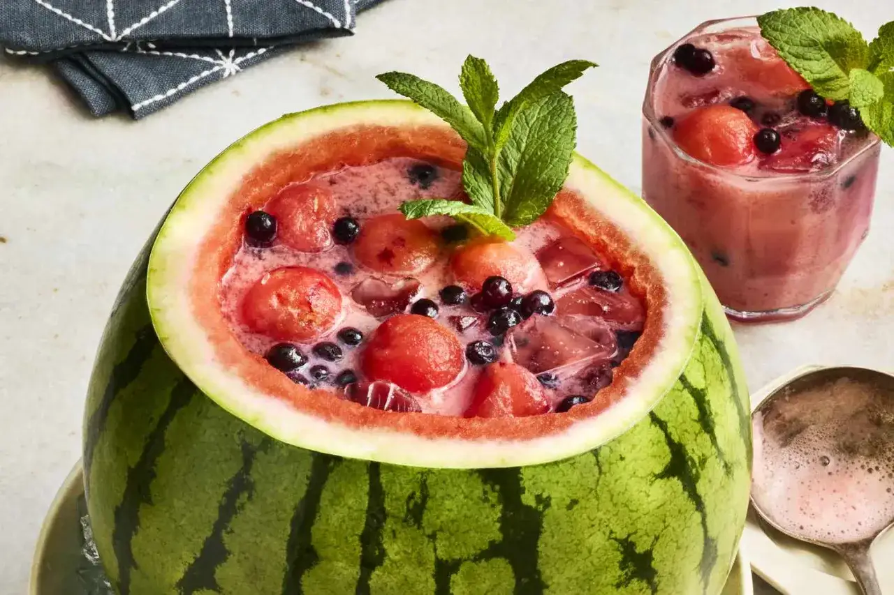 Orzeźwiający drink w arbuzie z kawałkami owoców i borówkami, ozdobiony miętą. Obok szklanka z podobnym napojem.