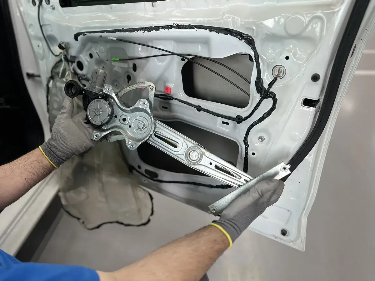 Mec&aacute;nico con guantes grises manipula el mecanismo del elevalunas no funciona, dentro de la puerta de un coche blanco.