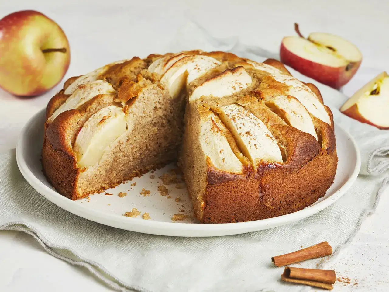 Ein saftiger Apfelkuchen ohne Zucker, garniert mit Apfelscheiben und Zimt. Daneben liegen frische Äpfel und Zimtstangen.