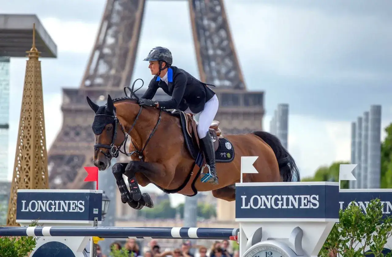 Cavalier et cheval brun sautent un obstacle devant la Tour Eiffel. La **flora de mariposa** s'épanouit dans ce décor parisien.