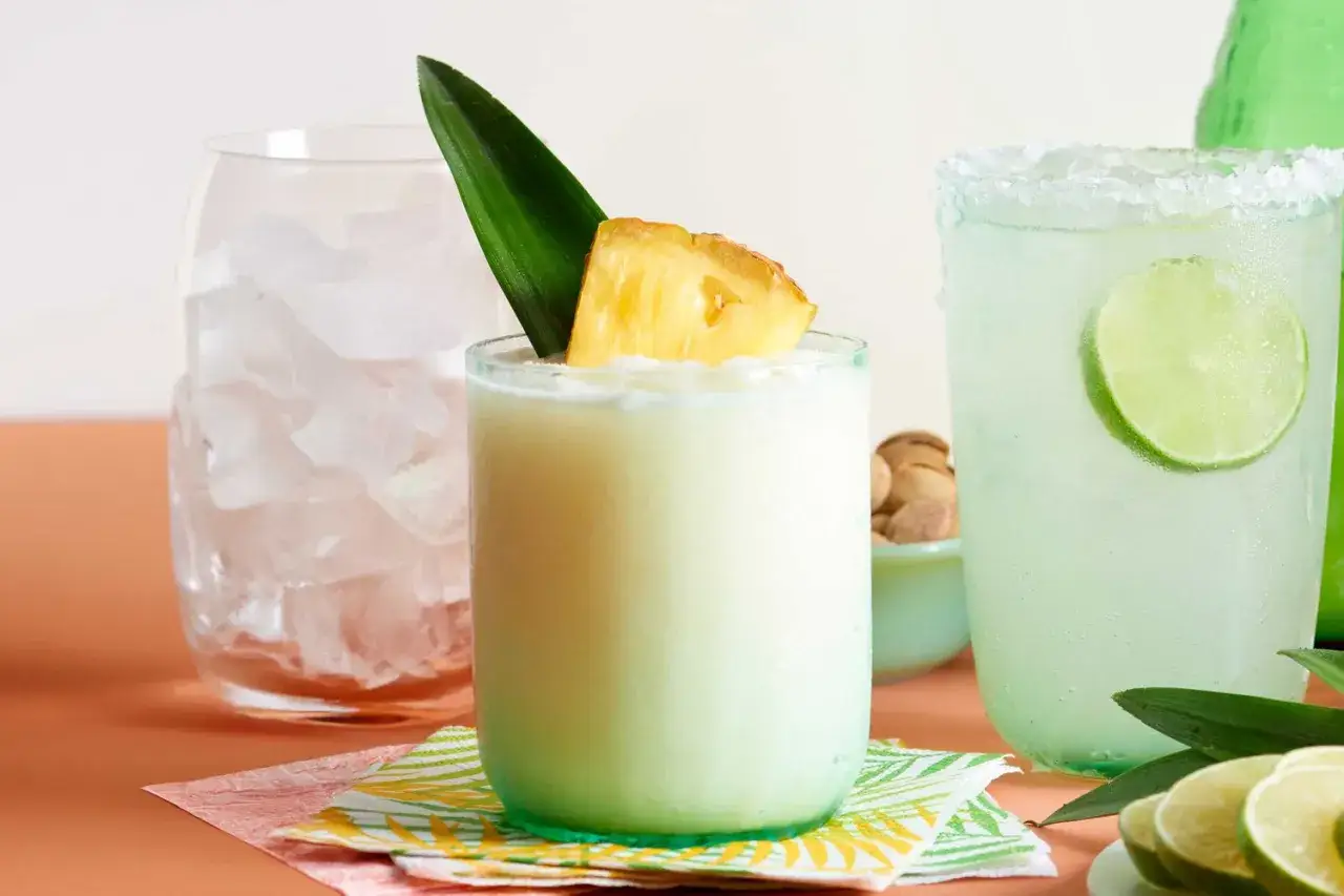 Pyszna pina colada, idealna na upalne dni, z kawałkiem ananasa i liściem. Obok drink z limonką.