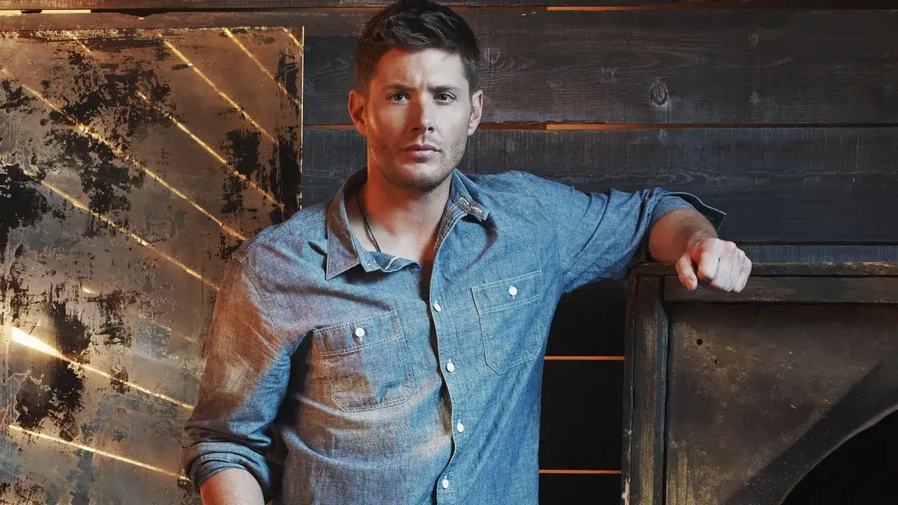 Jensen Ackles w dżinsowej koszuli, znany z seriali i programów, opiera się o drewnianą ścianę.