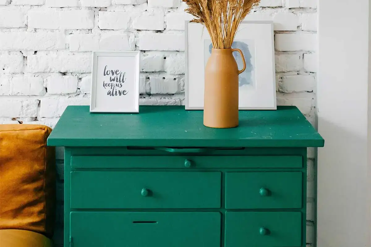 Un mobile in legno verde smeraldo, pronto per essere verniciato, con cornici e un vaso di fiori secchi.
