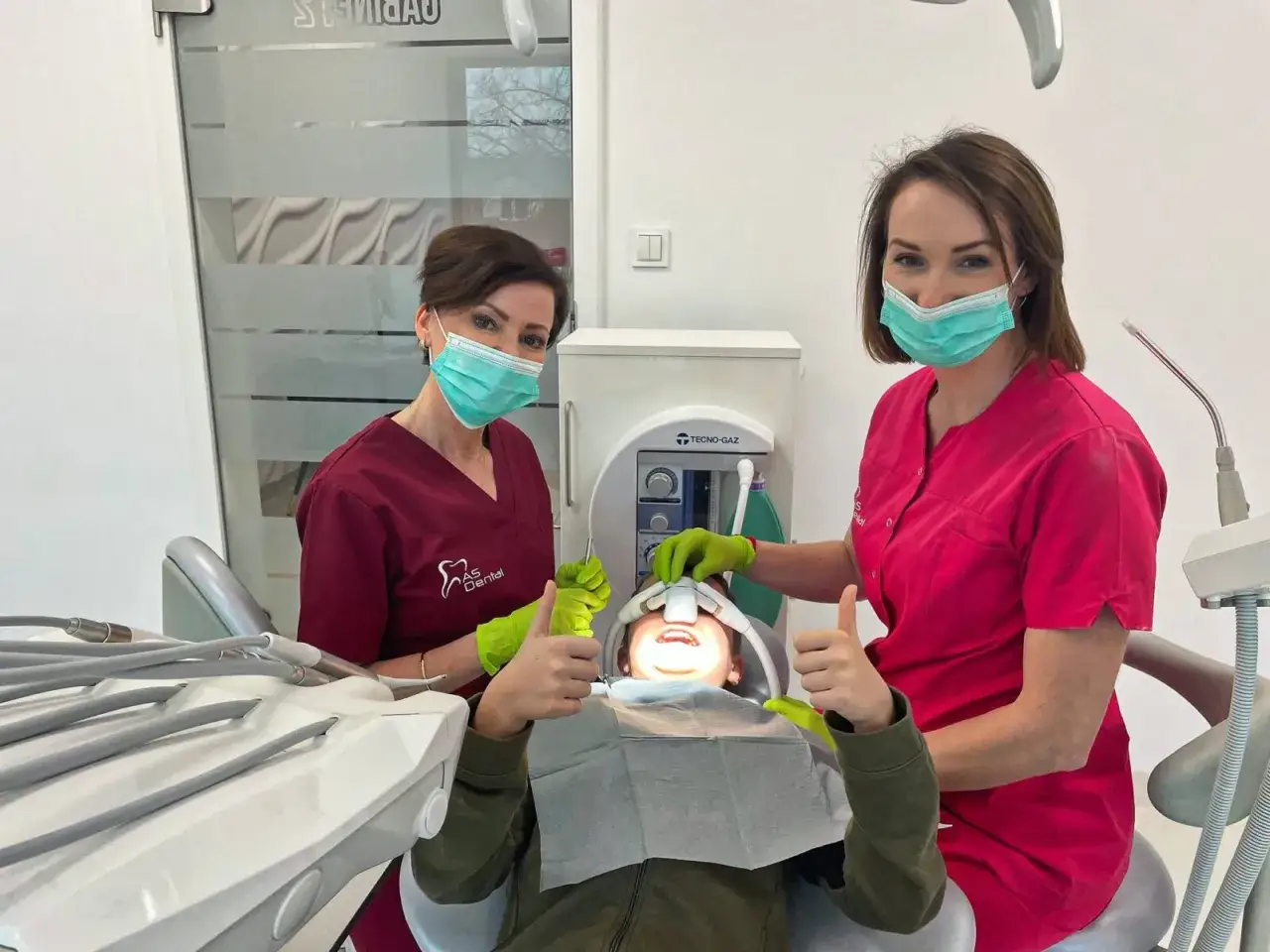 Dwie dentystki w maskach i rękawiczkach z uśmiechniętym pacjentem w gabinecie stomatologicznym.